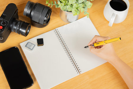 A person writing in a notebook with camera, memory cards, phone and lens on wooden table. concept of simple creative work spaceの写真素材