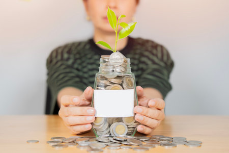A young woman sits at a table, holding a glass jar with green plant growth in coins. Selective focus, copy space.の写真素材