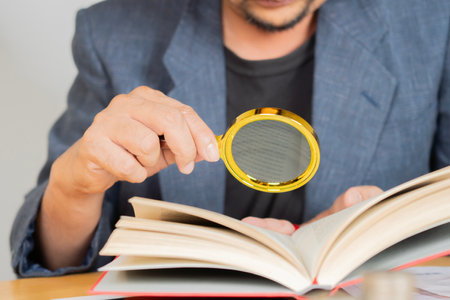 Businessman looking through a magnifying glass to a book on wooden table. Selective focus.の写真素材