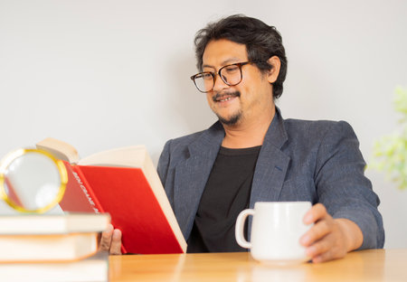 Handsome man with a beard and coffee reading a book sitting at desk in the living room. Education and success concept.の写真素材