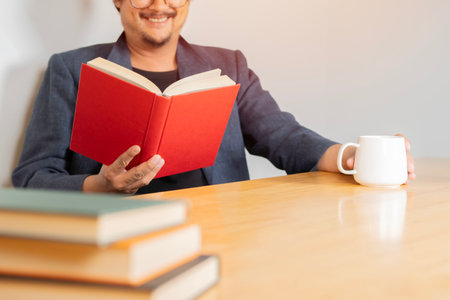Happy Asian businessman in casual suit, holding coffee cup and reading a book while sitting in reading room. Selective focus.の写真素材