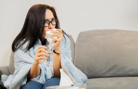 Asian woman cover with a blanket coughing and has a sore throat drinking water to relieve a sore throat in livingroom at home. Copy space.の写真素材