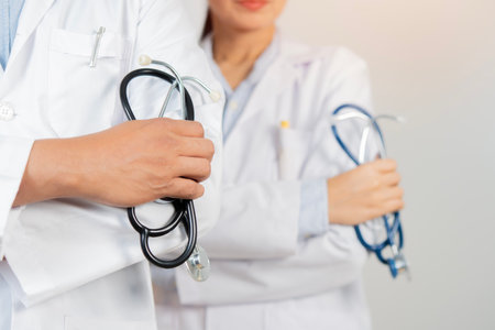 Cropped image of two determined physicians wearing white medical gowns standing and holding a stethoscope over white background. Selective focus.の写真素材