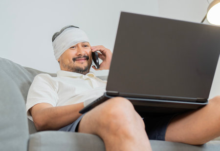 Bearded man with head injury sitting on sofa, uses laptop and phone at home for working online. Health care concept.の写真素材
