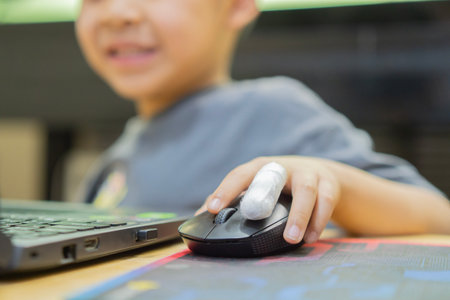 Smiling boy with bandaged on finger holding computer mouse and using laptop for play game or...