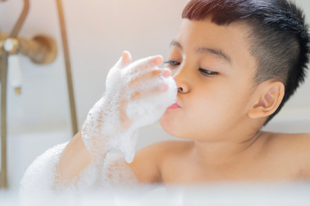 Happy little cute Asian boy bathes in a bath with foam and soap bubbles and playing and enjoying in...