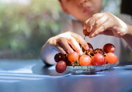 Happy child boy holding fresh red grapes with joyful expression at home with copy space. Kids Loves...