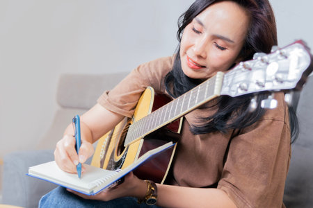 Happy Asian musician writing songs or taking notes while plays acoustic guitar at home. Songwriter concept.の写真素材