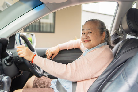 Smiling Asian senior woman driving car looking to camera while enjoying freedom and journey, hands...