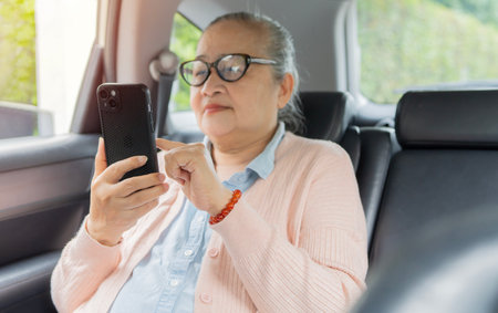 Senior Asian woman using her smart phone while sitting on the back seat in the modern car. Retirement lifestyle concept.の写真素材