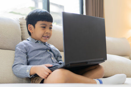 Happy Asian little cute boy sitting on sofa using laptop in online class at home. education, childhood, online learning, communication and domestic life, unaltered.の写真素材