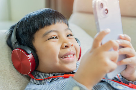 Cute little Asian boy with headphones and smartphone listening to audiobook at home. Close-up,...