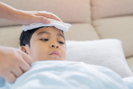 Little boy gets high fever lying on bed with cold compress, wet washcloth on forehead by his mom holding. Selective focus. Copy space.の写真素材