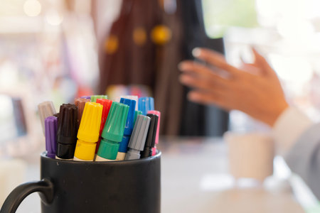 Multicolored marker pens in a black ceramic mug with blurred image of woman in an art workshop background. Selective focus.の写真素材