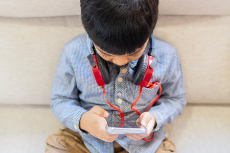 High angle view of young boy with headphone sitting on sofa, playing mobile games or watching...