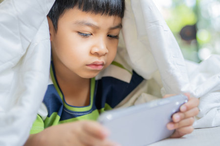 Small Asian little kid watching mobile phone at bed, blanket over his head, phone addiction. Selective focus.の写真素材