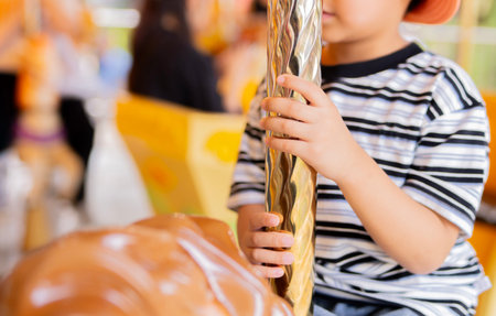 Close up of child hand with sstriped t-shirt holding golden spiral pole of horse at carousel in amusement park. Selective focus.の写真素材