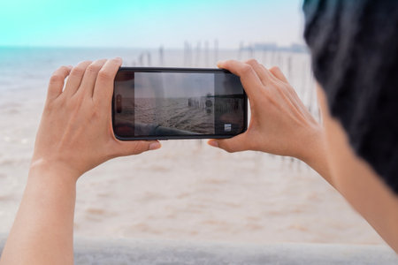 Rear view of woman in black wool hat taking picture with smartphone near sea on sunny day. Summer vacation, travel and technology conceptの写真素材