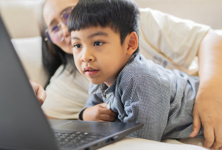 Smiling Asian mother and cute little son lying down on sofa while using laptop and looking at screen.の写真素材