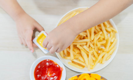 Little kid hands taking French fries from white plate with cheese dip, boy eating French fries in fast food cafe. Top view, selective focus.の写真素材