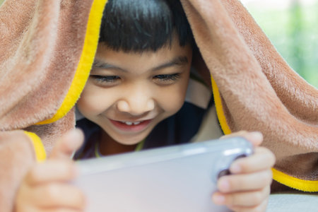 Happy cute little Asian boy with smartphone hiding under the brown blanket lying on bed at home, child watching cartoons or playing mobile games on phoneの写真素材