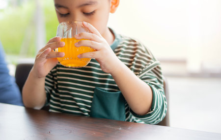 Little Asian boy sitting at an outdoor dining table and drinking orange juice at breakfast....