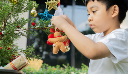 Holiday concept. Side view of Asian little boy prepare to decorate the Christmas tree, as a focused boy arranges beautiful ornaments and festive hifts at outdoor.の写真素材