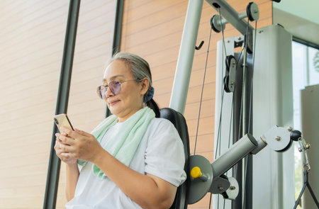 Active middle-aged Asian woman using smart phone while training at gym. Retirement lifestyle...