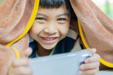 Cute little Asian boy using modern mobile phone under blanket at home.の写真素材