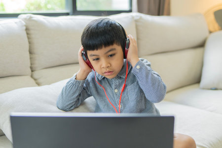 Asian boy sitting on sofa using laptop and headphones in online class at home. Selective focus.の写真素材