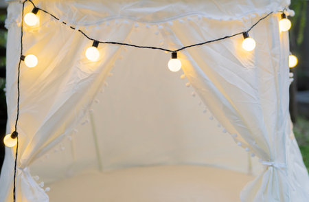Emtpy of kids room in light colors with teepee tent and bulbs. Cozy tent with fairy lights creating...