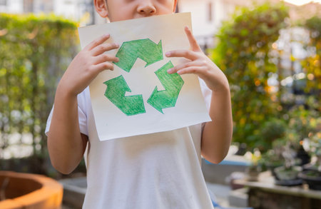 Little child boy standing and holding in hands recycling sign symbol over home garden background. Ecological sorting garbage concept.の写真素材