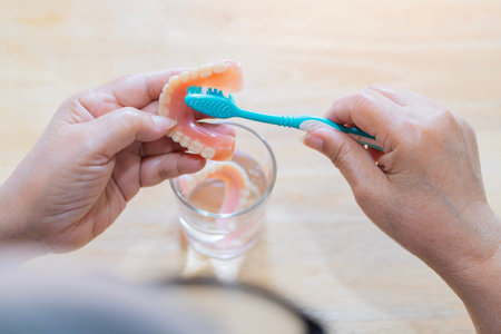 Rear view of an elderly person holds a full upper denture while cleaning it with a toothbrush on table. illustrating proper hygiene care.の写真素材