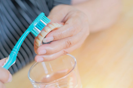 An elderly person scrubs a denture with a toothbrush and glass of water on table, demonstrating proper denture cleaning and oral hygiene maintenance. Selective focus, copy space.の写真素材