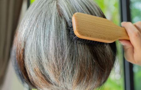 Elderly woman with gray hair combing with wooden comb for healthy hair. Care Routine concept. Back...