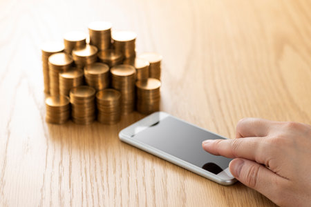 Smartphone with coins stacked on wooden table.の写真素材