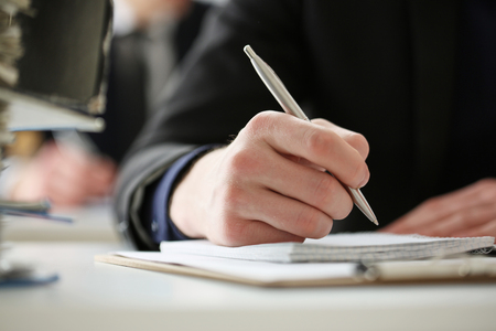 Male hand holding silver pen ready to make note in opened notebook sheet. Businessman in suit at workspace make thoughts records at personal organizer, white collar conference, signature conceptの写真素材