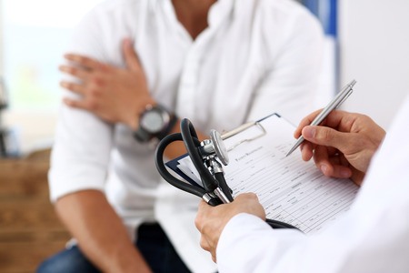 Male doctor hold silver pen filling patient history list at clipboard pad. Physical, exam, er, trauma pain, disease prevention, ward round, 911, prescribe remedy, healthy lifestyle conceptの写真素材