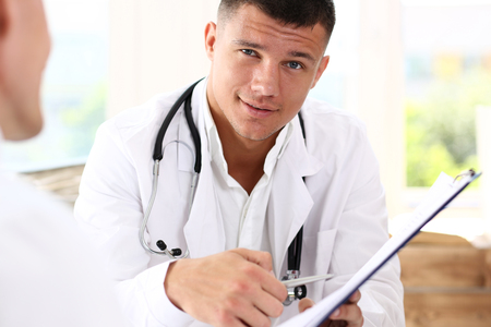 Smiling handsome doctor communicate with patient holding silver pen and showing pad. Physical agreement signature, disease prevention, consent sign, 911, prescribe remedy, healthy lifestyle conceptの写真素材