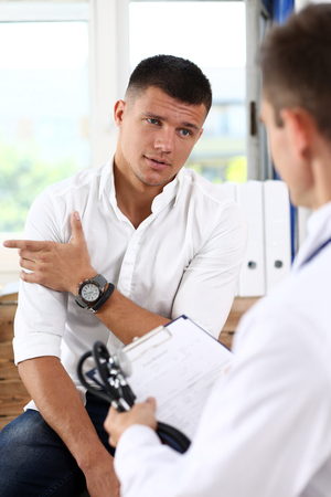 Male patient portrait with doctor filling patient history list at clipboard pad. Physical, exam, er, trauma pain, disease prevention, ward round, 911, prescribe remedy, healthy lifestyle conceptの写真素材