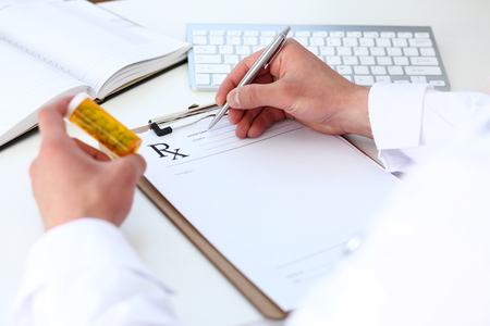 Male medicine doctor hand hold jar of pills and write prescription to patient at worktable. Panacea and life save, prescribing treatment, legal drug store concept. Empty form ready to be usedの写真素材