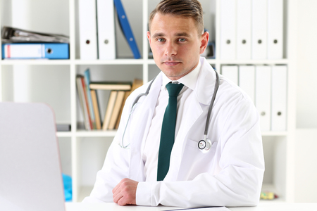 Handsome smiling male doctor write sit at workplace in office portrait. Panacea and life save, reception physical, prescribing treatment, legal drug store, take stock, consumption statistics conceptの写真素材