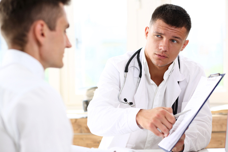 Concerned handsome doctor communicate with patient holding silver pen and showing pad. Physical agreement signature, disease prevention, consent sign, 911, prescribe remedy, healthy lifestyle conceptの写真素材
