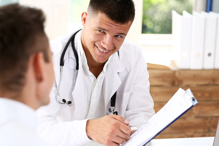 Smiling handsome doctor communicate with patient holding silver pen and showing pad. Physical agreement signature, disease prevention, consent sign, 911, prescribe remedy, healthy lifestyle conceptの写真素材