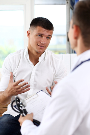 Male patient portrait with doctor filling patient history list at clipboard pad. Physical, exam, er, trauma pain, disease prevention, ward round, 911, prescribe remedy, healthy lifestyle conceptの写真素材