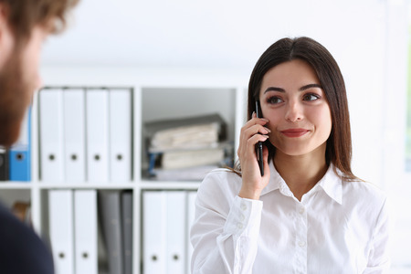 Beautiful businesswoman brunette portrait in an office in a white shirt is holding a phone and talking to a customer remotely looking into the distance listening to what they say and receiving information remotelyの写真素材