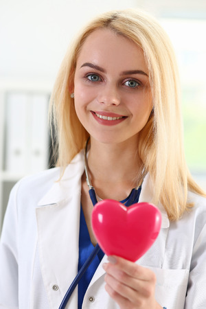 Female medicine doctor hold in hands red toy heart in front of her chest. Cardio therapeutist, student education, physician make cardiac physical, heart rate measure, arrhythmia conceptの写真素材