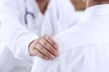 Friendly male doctor hold patient shoulder in office during reception. Examination result, positive test, calm down, promise and cheer up, grief and suffer, treatment, condolence, ethics conceptの写真素材