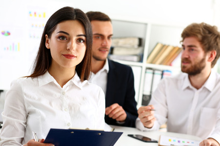 Beautiful smiling cheerful girl at workplace look in camera with colleagues group in background. White collar worker at workspace, job offer, modern lifestyle, client visit, profession train conceptの写真素材