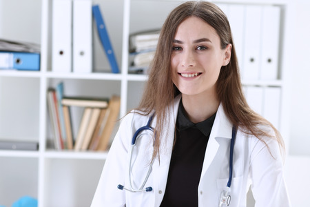 Beautiful smiling female doctor sit at workplace. Physical and disease prevention, patient aid, exam visit, 911, ward round, prescribe remedy, healthy lifestyle, consultant profession conceptの写真素材